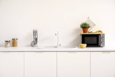 Beyaz tezgahlarda mikrodalga fırın, lavabo ve mutfak aletleri olan bir mutfak.