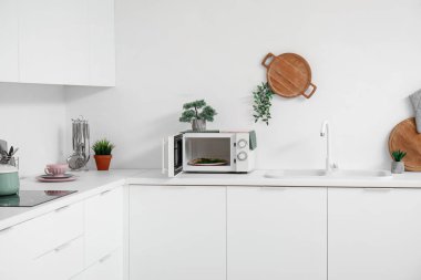 Beyaz tezgahlarda mikrodalga fırın, lavabo ve mutfak aletleri olan bir mutfak.