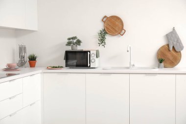 Beyaz tezgahlarda mikrodalga fırın, lavabo ve mutfak aletleri olan bir mutfak.