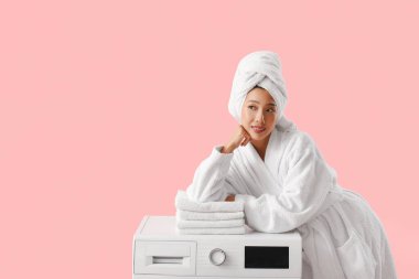 Bornozlu güzel bir kadın çamaşır makinesinin üzerinde pembe arka planda bir yığın temiz kıyafetle.