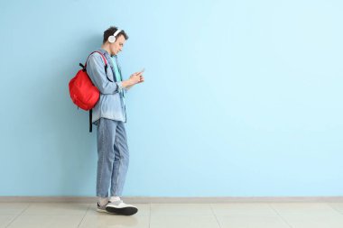 Sırt çantalı ve cep telefonlu bir erkek öğrenci mavi duvarın yanında kulaklıkla müzik dinliyor.