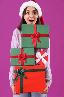 Mor arka planda hediye kutuları olan Noel Baba şapkalı mutlu bir genç kadın.
