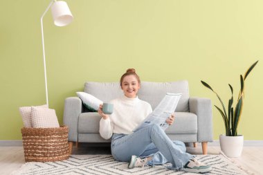 Elinde kahve fincanı olan genç bir kadın evde gazete okuyor.