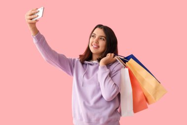 Genç bir kadın selfie çekiyor ve pembe arka planda alışveriş torbaları tutuyor.