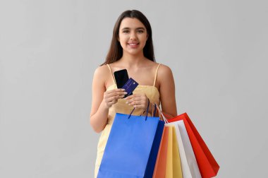 Gri arka planda kredi kartı, cep telefonu ve alışveriş çantaları olan genç bir kadın.