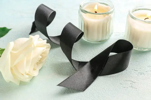 Beautiful rose flower, burning candles and black funeral ribbon on blue background