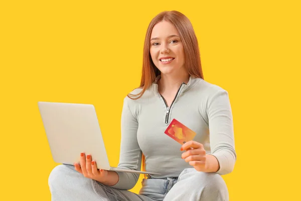 Young woman with credit card and laptop sitting on yellow background