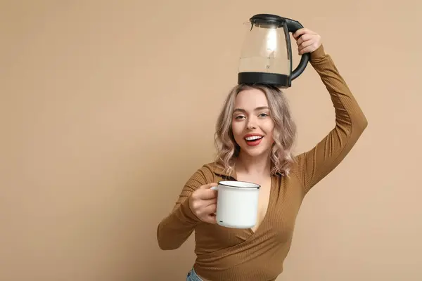 Funny young woman with modern electric kettle and mug on beige background