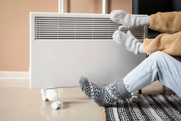 Evde elektrikli ısıtıcının yanında sıcak çorap ve eldiven giyen genç bir kadın var. Isınma mevsimi kavramı