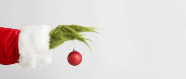 Green hairy hand of creature in Santa costume holding Christmas ball on light background with space for text clipart