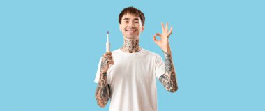 Elektrikli diş fırçası olan genç dövmeli adam açık mavi arka planda iyi görünüyor.