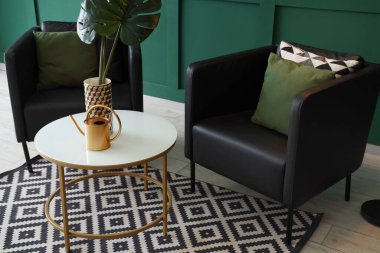 Yastıklı siyah modern koltuklar, masa ve lamba yeşil duvarın yanında.