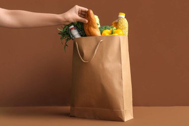 Kağıt alışveriş torbası ve kahverengi arka planda farklı taze ürünleri olan bir kadın.