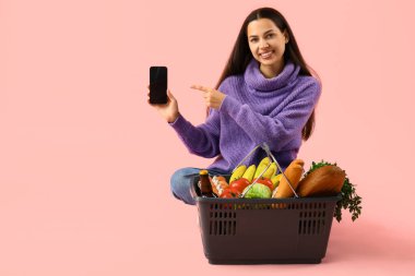 Elinde alışveriş sepeti olan genç bir kadın pembe arka planda cep telefonuna bakıyor.