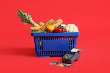 Ödeme terminali ve kredi kartlarıyla dolu alışveriş sepeti kırmızı arka planda.