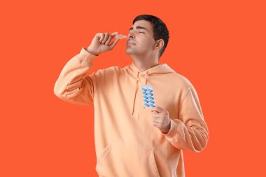 Turuncu arka planda burun damlası kullanan haplı genç bir adam.