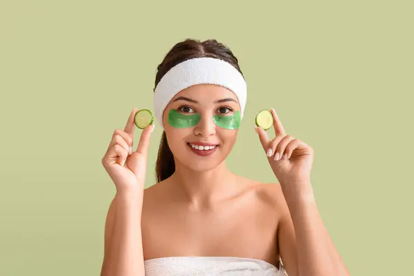 Pretty young woman with under-eye patches and cucumber slices on green background