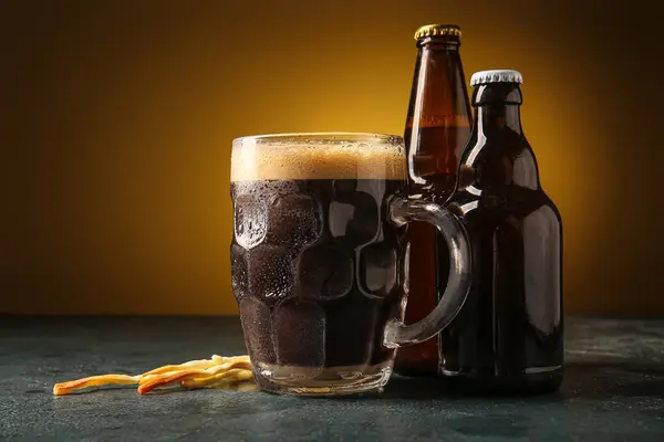 Bottles and mug of dark beer with cheese on table against yellow background