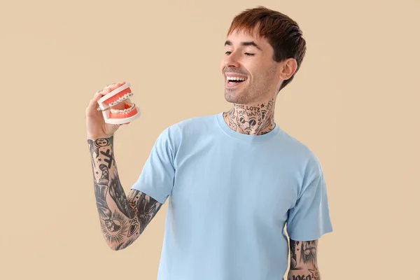 Young tattooed man holding jaw model with braces on beige background