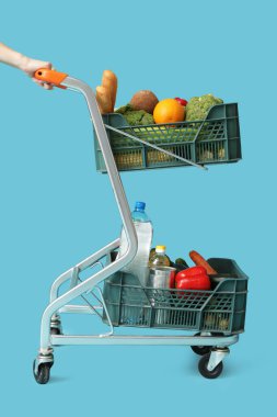 Mavi arka planda yiyecek dolu alışveriş arabasını iten kadın.