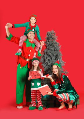Mutlu aile elf kostümleri içinde hediye kutuları, Noel Baba kartları ve kırmızı arka planda Noel ağacı.