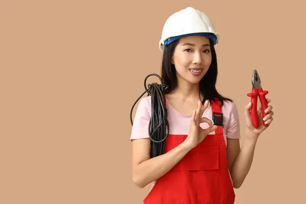 Young female electrician with pliers and cables showing OK gesture on beige background