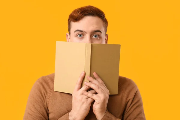 Young man covering his face with book on yellow background, closeup
