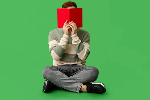 Young man covering his face with book on green background