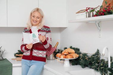 Mutfakta Noel kutlaması için tebrik kartı taşıyan olgun bir kadın.