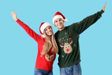 Mavi arka planda Noel Baba şapkalı ve Noel kazaklı genç bir çift.