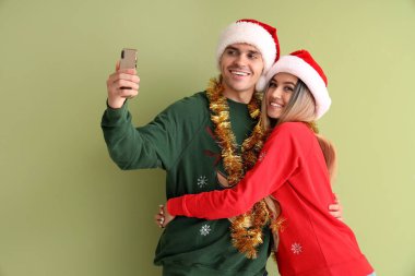 Noel Baba şapkalı ve Noel kazaklı genç çift yeşil arka planda selfie çekiyorlar.