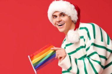 Kırmızı arka planda LGBT bayrağı olan Noel Baba şapkalı genç adam.