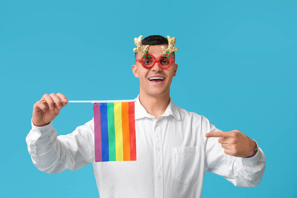 Young man in Christmas eyeglasses pointing at LGBT flag on blue background