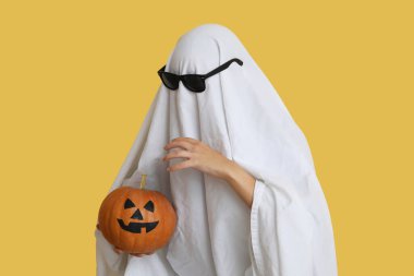 Cadılar Bayramı kostümlü kadın hayalet ve güneş gözlüğü renkli arka planda balkabağı tutuyor.