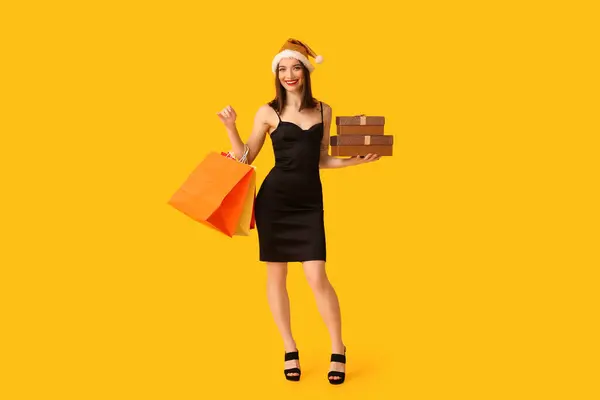 黄色の背景にショッピングバッグとクリスマスギフトボックスとサンタハットの若い女性 — ストック写真