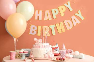 Koyu bej duvarın yanındaki beyaz masada mumlar ve şekerler olan doğum günü pastası.