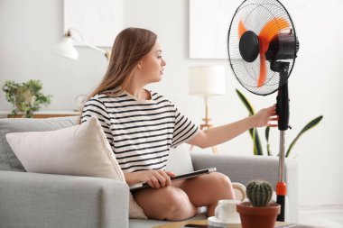 Evde dizüstü bilgisayarı ve elektrikli vantilatörü olan genç bir kadın var.