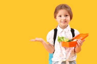 Sırt çantalı ve beslenme çantalı mutlu küçük kız sarı arka plandaki bir şeyi işaret ediyor.