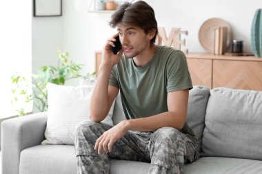 Genç erkek asker evdeki koltukta otururken cep telefonuyla konuşuyor.