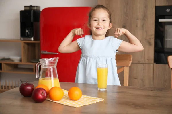 Little girl with glass of orange juice showing muscles in kitchen