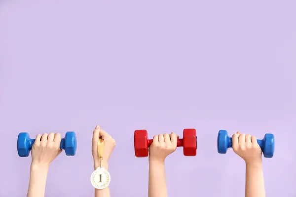 Female hands with dumbbells and medal on lilac background