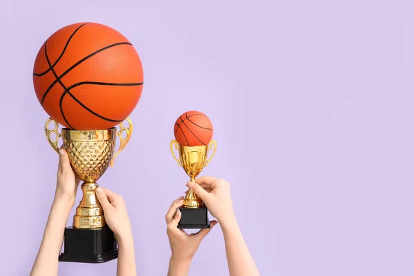 Female hands with gold cups and basketballs on lilac background