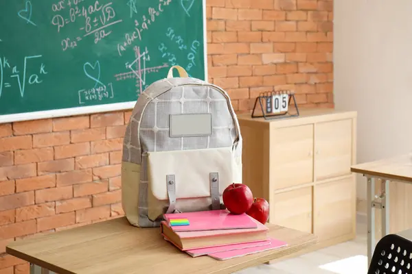 Stylish school backpack with stationery and fresh apples on desk in classroom