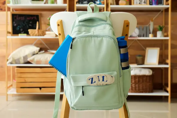 Stylish school backpack with bottle of water and stationery hanging on chair in room