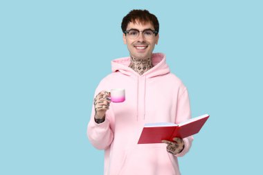 Mavi arka planda kitap ve bir fincan kahve olan dövmeli genç bir adam.