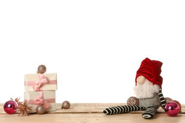 Kahverengi ahşap masada beyaz arka planda hediye kutusu ve topları olan Noel cücesi.