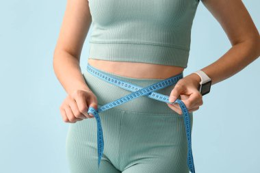 Beautiful young woman with measuring tape on blue background, closeup. Weight loss concept clipart