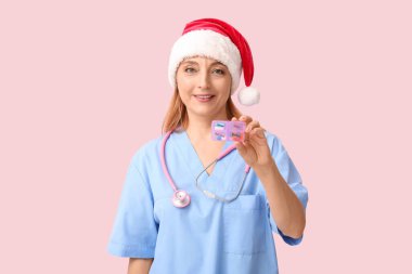 Pembe arka planda haplarla dolu Noel Baba şapkalı olgun bir kadın doktor.