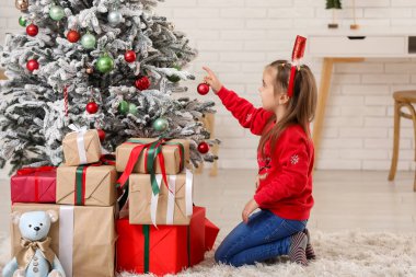 Noel Baba şapkalı şirin bir kız Noel ağacını süslüyor ve evde hediye kutuları var.