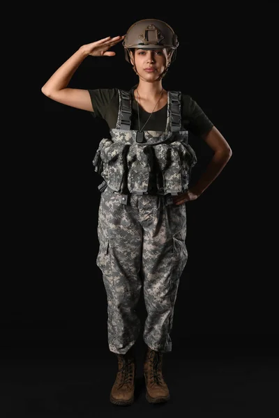 Confident young woman in military uniform saluting on black background